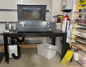 This image shows a home laser cutting setup. On a sturdy black table sits a large laser cutter machine, the xTool P2, with a clear window for viewing the cutting process. Beneath the table, there is a connected air filter system with black vent hoses, used to safely filter fumes. To the right, a yellow duster, cleaning supplies, and storage containers are neatly placed, contributing to a clean, organized workspace. laser cutting