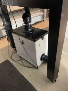 The image shows an air filtration unit placed beneath a sturdy black table. The unit is white with a black top and is connected to a flexible black hose that extends upward. The hose appears to lead to a laser cutter (not visible in this specific image). There are electrical cords connected to the filtration unit, and it is positioned on a smooth concrete floor. This air filtration system is likely used to extract fumes and particles generated during the laser cutting process, ensuring a cleaner and safer workspace.