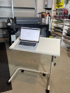 The image shows a small, height-adjustable white table on wheels with a silver metal frame. On top of the table sits an open laptop displaying a design application. In the background, there is a larger workspace, including a laser cutter and various tools and supplies, neatly organized on shelves. The table is positioned near the laser cutter, indicating that the laptop is likely used for controlling or designing laser-cutting projects. The space appears clean and well-organized for crafting or making.