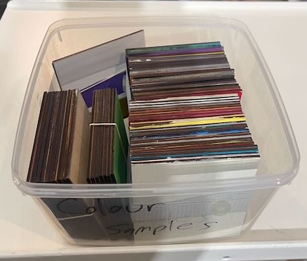 The image shows a plastic container filled with various color samples of materials, likely used for laser-cutting or other crafting projects. Each sample appears to be a small rectangular or square piece, stacked neatly, showcasing a range of colors and finishes. The variety of samples suggests that this box is a go-to resource for anyone looking to choose the right material or color for their next project, offering a broad selection of potential materials for creative endeavors.