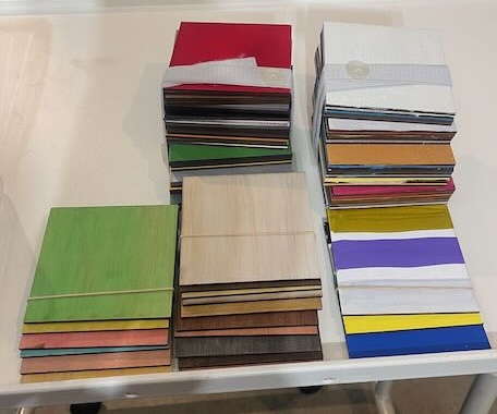 Three stacks of flat, square, colored acrylic samples are arranged on a table. Each stack contains sheets of varying colors and finishes. The colors range from natural wood grains to bold shades like green, yellow, red, and purple. These materials are typically used in laser cutting projects to help determine the right color and finish for a design.