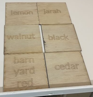 This image depicts six square wooden tiles, each engraved with the names of different wood colors. The colors represented include lemon, jarah, walnut, black, barn yard red, and cedar. The text on each tile is lightly etched into the wood, with the wood grain still visible across the tiles. These tiles are likely samples showing different wood finishes.