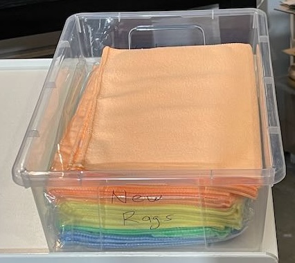 A clear plastic container holds neatly folded, colorful microfiber cloths labeled as "New Rags." The cloths are arranged in a visible spectrum of colors including orange, green, blue, and yellow, showing they are ready for use. The transparent bin allows easy access and organization, ensuring these cloths are stored in a clean, dust-free environment. Suitable for various tasks, such as wiping, cleaning, or polishing, these rags are versatile for household or workshop use, maintaining a fresh and tidy appearance.