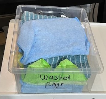 This image shows a clear plastic storage bin labeled "Washed Rags." Inside the bin, there are neatly folded cleaning rags, including light blue microfiber cloths and a couple of green rags, stacked on top of each other. The blue rags have a smooth, soft texture, while the green ones appear more textured, suitable for cleaning various surfaces. The bin is transparent, making the contents visible, and it sits on a white surface in a workspace or storage area.