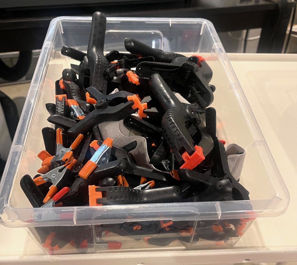 This image shows a clear plastic container filled with multiple black and orange plastic clamps. The clamps have orange gripping ends and black handles, designed for easy one-handed use. These spring-loaded clamps are commonly used to hold materials in place while glue dries or to secure items during projects. The clamps vary slightly in size, and the bright orange parts stand out against the black handles, making them easily visible. The container is filled to the top, ensuring an ample supply for various crafting or woodworking tasks.