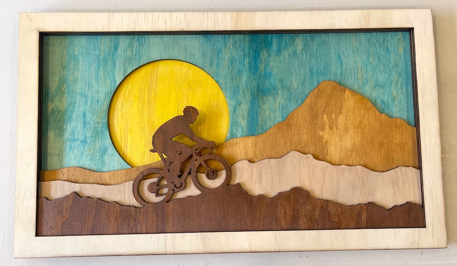 Sunset Ride: Laser-Cut Mountain Biking Adventure Laser Cutting Project - This laser-cut wall art captures the essence of a mountain biking adventure against a vibrant backdrop. Featuring a cyclist riding across layered mountain terrains, the piece is highlighted by a warm, glowing sun setting against a turquoise sky. The multi-layered design adds depth and dimension, creating a stunning, dynamic scene that celebrates the thrill of outdoor biking. Perfect for cycling enthusiasts, this artwork brings a sense of adventure and natural beauty to any space.