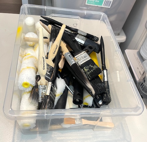 A clear plastic container filled with different paintbrushes and small paint rollers, featuring brushes with wooden and black handles, suitable for detailed painting and larger projects.