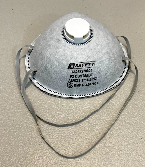 This image shows a standard P2 dust and mist respirator mask with a white, dome-shaped design and elastic bands. The mask has a central valve for easier breathing and is labeled with safety standards, indicating it provides protection against dust and mist particles. The mask is intended for use in environments where dust or particles may be present, providing essential respiratory protection for various tasks, such as sanding or laser cutting projects.