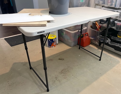 A foldable, lightweight work table stands open with a flat, sturdy plastic surface and black adjustable metal legs. On the table sits some plywood sheets and a grey bucket, while beneath it are various containers and tools, indicating it’s used for DIY or crafting projects. This versatile table provides a reliable workspace, adaptable for various tasks such as laser cutting, painting, or assembly, making it a valuable addition to any workshop or garage setup.