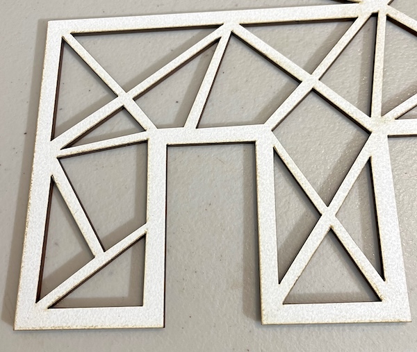 This image features a close-up view of a laser-cut design in white material. The cutout showcases a geometric pattern composed of interconnected triangles and rectangles, creating a modern and angular aesthetic. The edges of the cutouts have a slightly darkened tone, likely from the laser cutting process, adding subtle contrast against the white surface. The material appears smooth, with precise, clean cuts that highlight the accuracy of the laser-cutting technique.