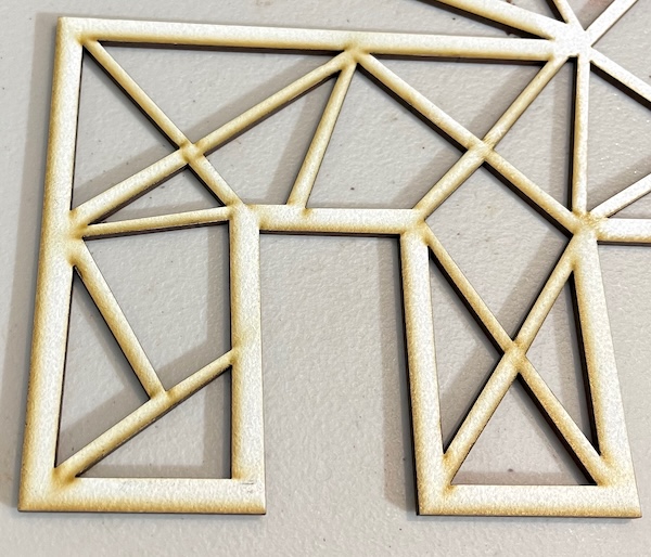 This image depicts a close-up of a laser-cut geometric design on a light-colored material. The pattern consists of intersecting triangles and rectangles, forming an angular, contemporary layout. The edges of the cutouts have a slightly charred or darkened appearance, a characteristic of the laser-cutting process. The surface shows some residual marks from the cutting, giving it a raw, unfinished look. The intricate design highlights the precision and detail achievable with laser-cutting technology.