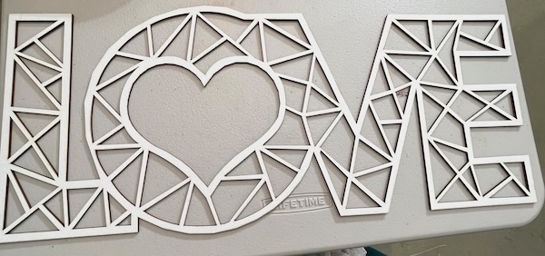 The image shows a laser-cut decorative design spelling the word "LOVE." The letters are composed of geometric patterns made up of triangles and rectangles, with a heart shape integrated into the letter "O." The material is a clean, light-colored surface, giving it a polished and refined look. The intricate design is symmetrical and visually striking, showcasing precision laser cutting and a modern, artistic style suitable for home decor or personalized gifts.