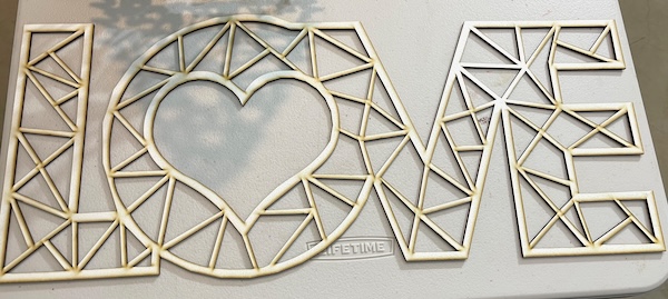 The image depicts a laser-cut decorative design spelling the word "LOVE," made from a light-colored material. The letters feature intricate geometric patterns of interconnected triangles and rectangles, with a heart shape integrated into the "O." The surface appears slightly unfinished, showing subtle scorch marks from the laser cutting process, indicating it is yet to be cleaned. This artistic and modern design demonstrates the precision and creativity of laser cutting, suitable for personalized gifts or decor.